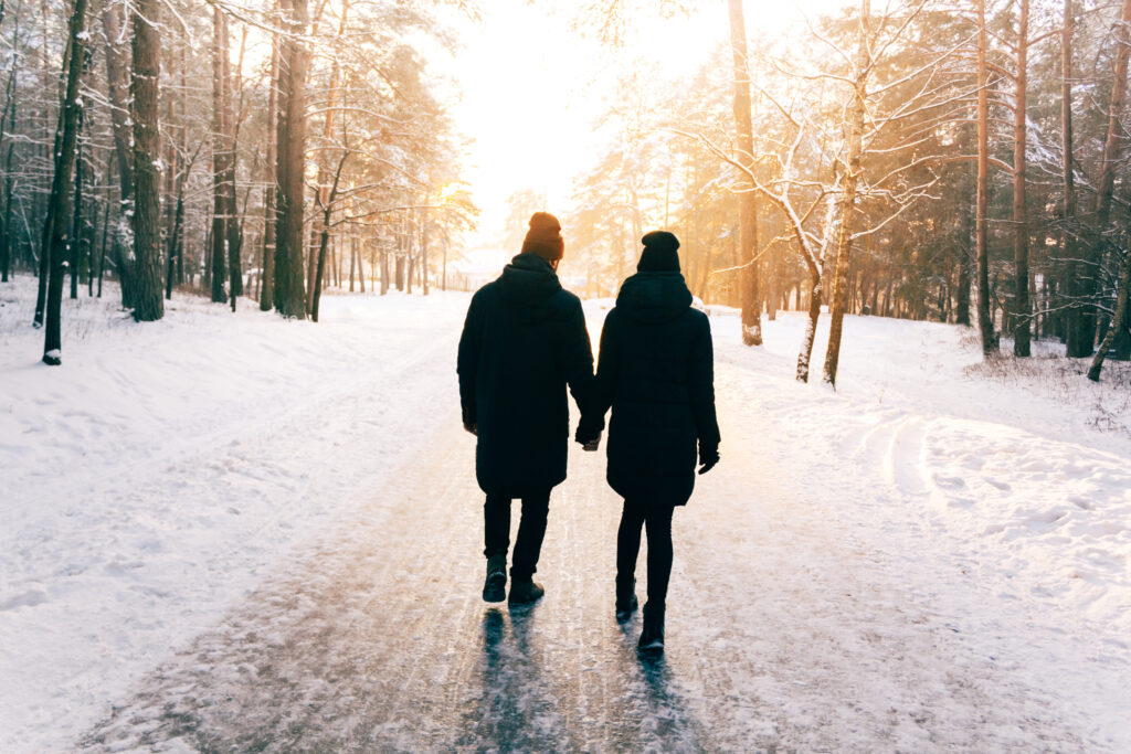 Two people walking down a snowy path in the forest hand-in-hand with their backs to the camera dressed in winter attire with the sun setting in front of them