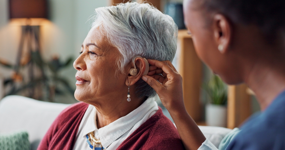 an older ethnic woman is seated while a nurse helps to fit a hearing aid into her ear.