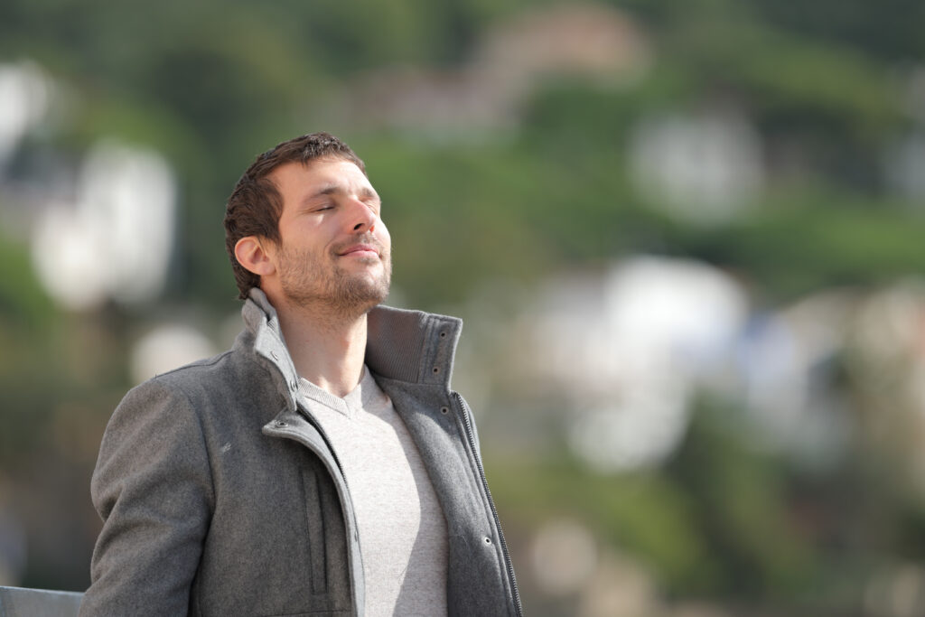 man in coat stands in the sun looking relaxed with tree background