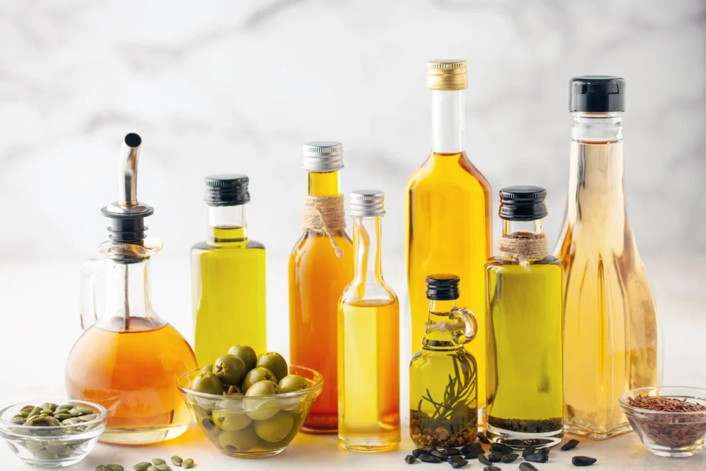 A variety of glass bottles filled with different cooking oils arranged on a light surface, with a small bowl of green olives, seeds, and herbs in front.