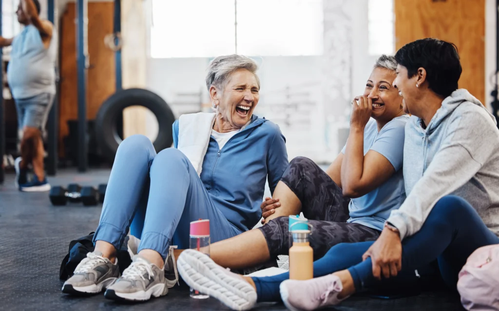 Three older women in workout clothes sit on a gym floor laughing together during a break, with water bottles beside them and exercise equipment in the background.
