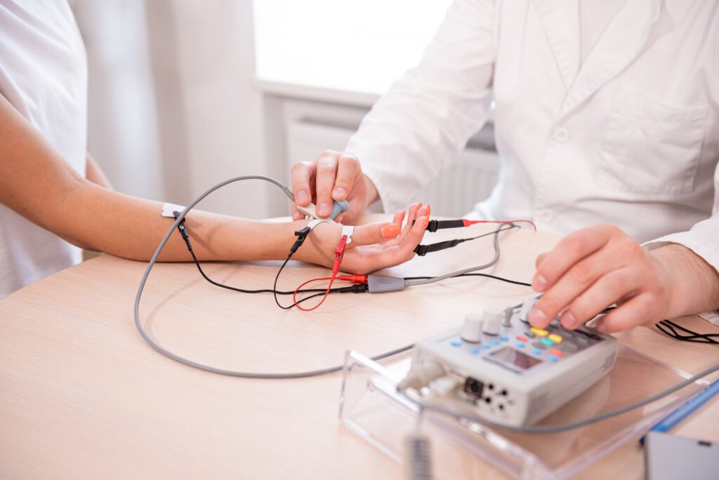 A patient and a doctor sit at a table in a white room. The patient's hand is resting on the table. There are red, black, and gray wires attached to the patients hand in different places. They are undergoing an electromyography test that measures muscle response to nerve stimulation.