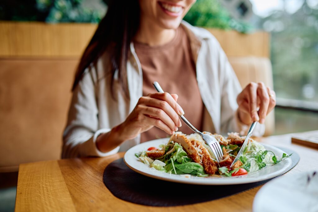 a smiling woman with brown hair in a tan shirt sits at a table eating a salad with fresh chicken.