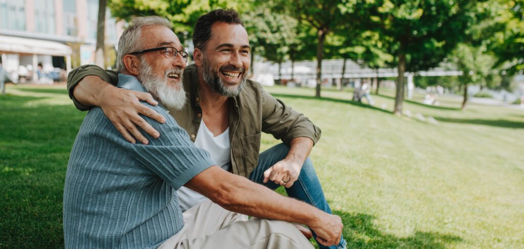 A young man sits with his father in a lush green park. Both of them are smiling.