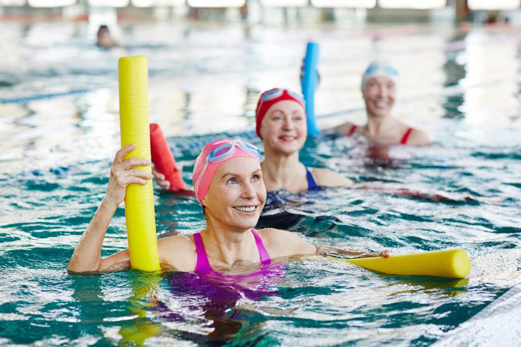 Three smiling women are floating in a public pool. They are doing water aerobics. They are wearing swimming caps and goggles while they balance on pool noodles.