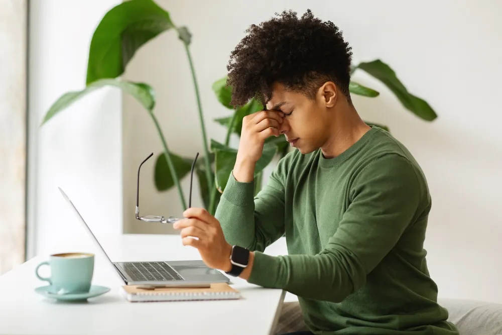 Person of color sits at a desk with computer. They have removed their glasses and are squeezing their brow uncomfortably.