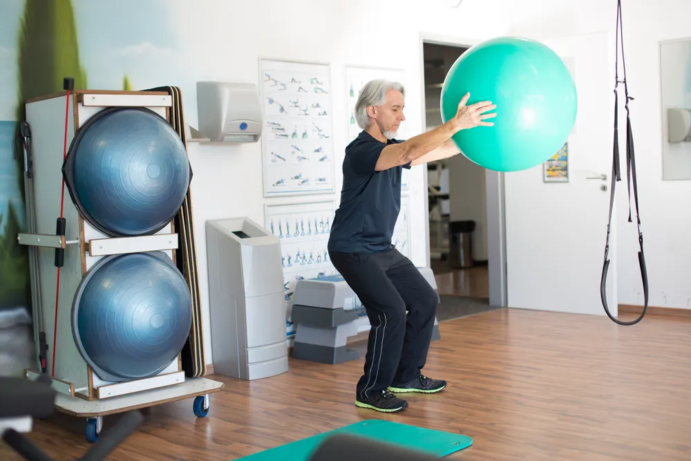 White older man works out in a room with various gym equipment. He is holding a medicine ball.