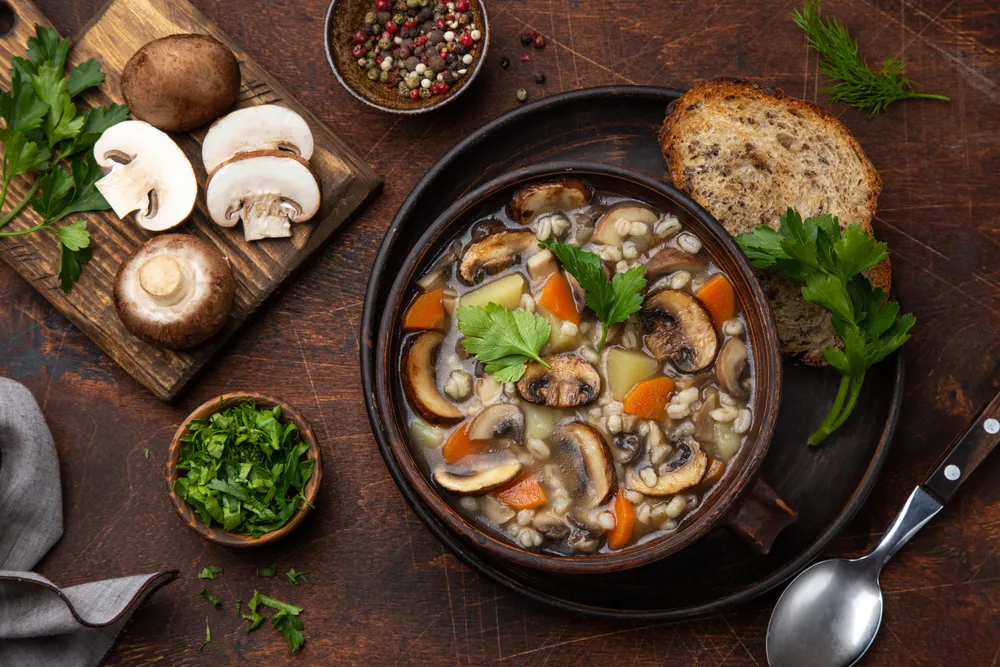 a bowl of mushroom barley soup sits on a wooden table. There are cut mushrooms, parsley, bread and spices surrounding the bowl.